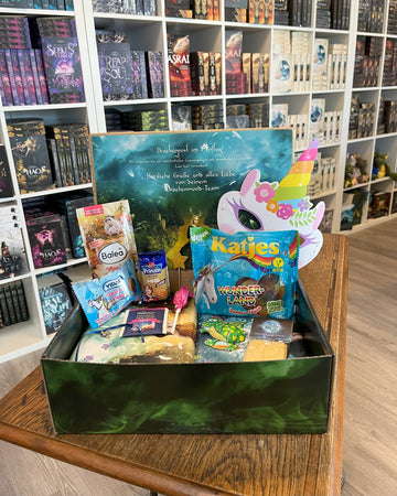 Das Produktbild zeigt vorn eine geöffnete Drachenpost in Märchen-Design. Man sieht den in den Boxdeckel geklebten Brief zum Thema Märchen und in der Box zum Thema passende Goodies - Süßigkeiten und andere Kleinigkeiten. Im Hintergrund sieht man die Bücherregale des Verlagsladens.